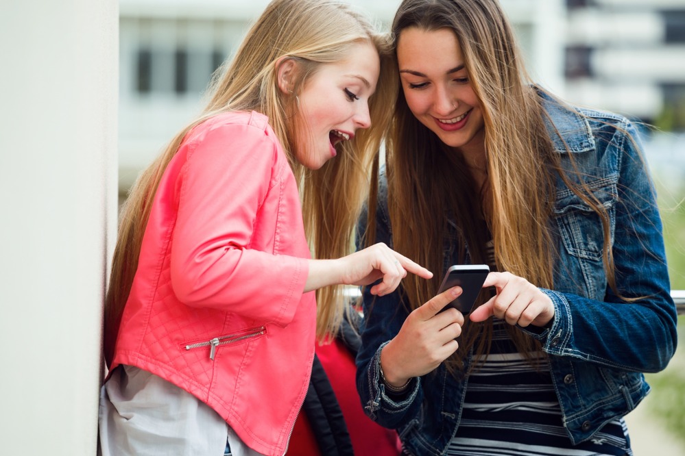 two-beautiful-students-using-mobile-phone-street.jpg